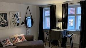 A modern living room with a brown sofa, a small round table, a dining area with a chair, and a window with curtains. - Apartment 1 bedroom Grønnegade (Copenhagen)