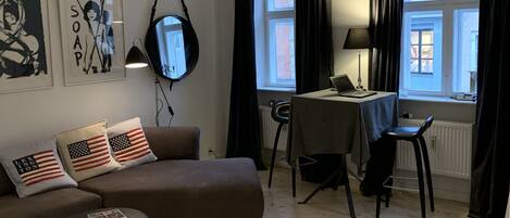 A modern living room with a brown sofa, a small round table, a dining area with a chair, and a window with curtains.
