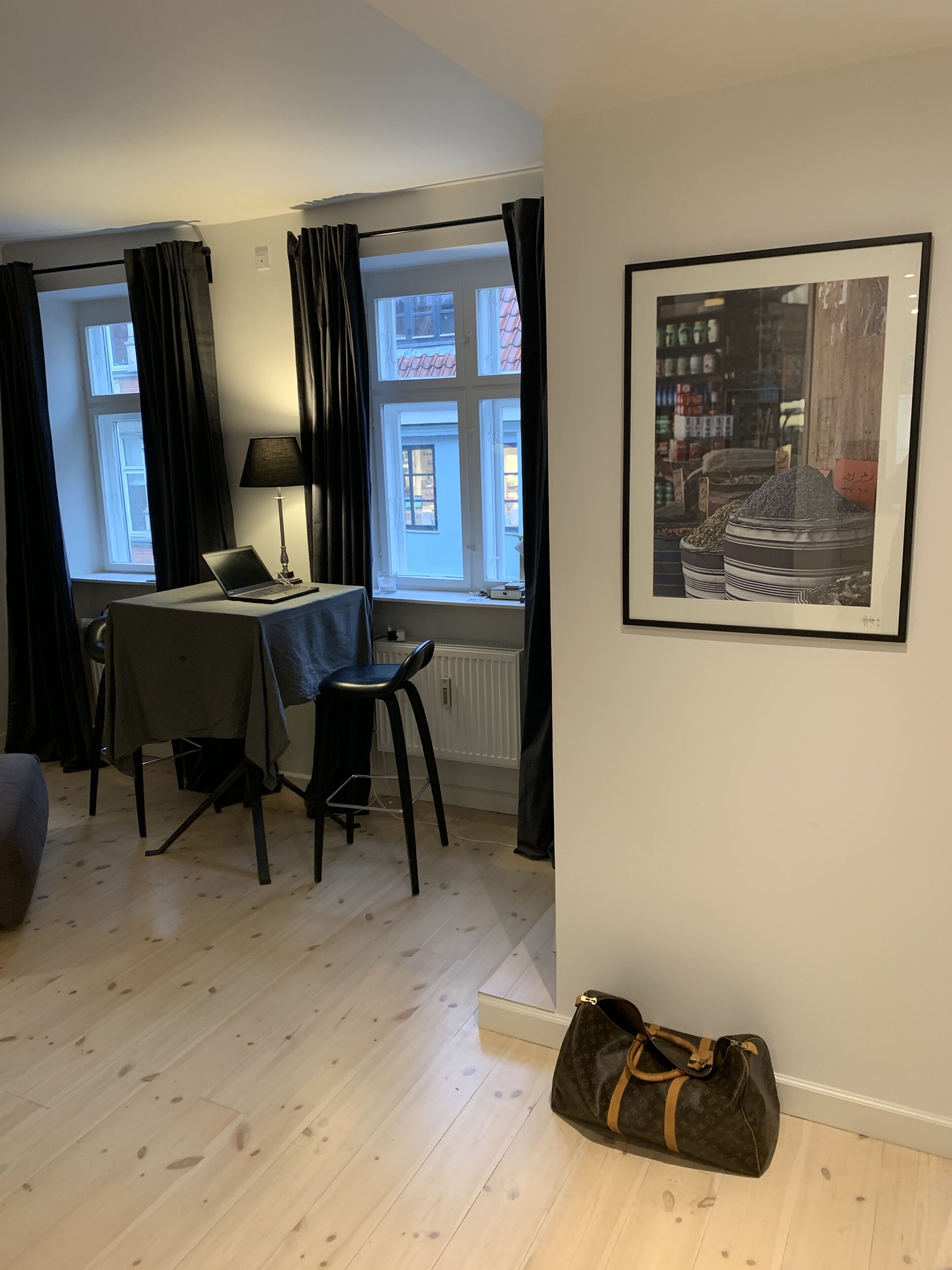 Ett rum med ett matbord, en bärbar dator, en stol, ett element, ett fönster med gardiner, en väska och en inramad tavla på väggen.