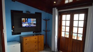 A room with a television mounted on the wall, a wooden cabinet, a small refrigerator, and a large wooden door with windows. - Pousada Souza Reis (Sao Thome das Letras)