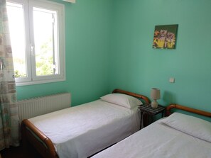 A room with two single beds, a window with a view of greenery, a radiator, and a small bedside table with a lamp.