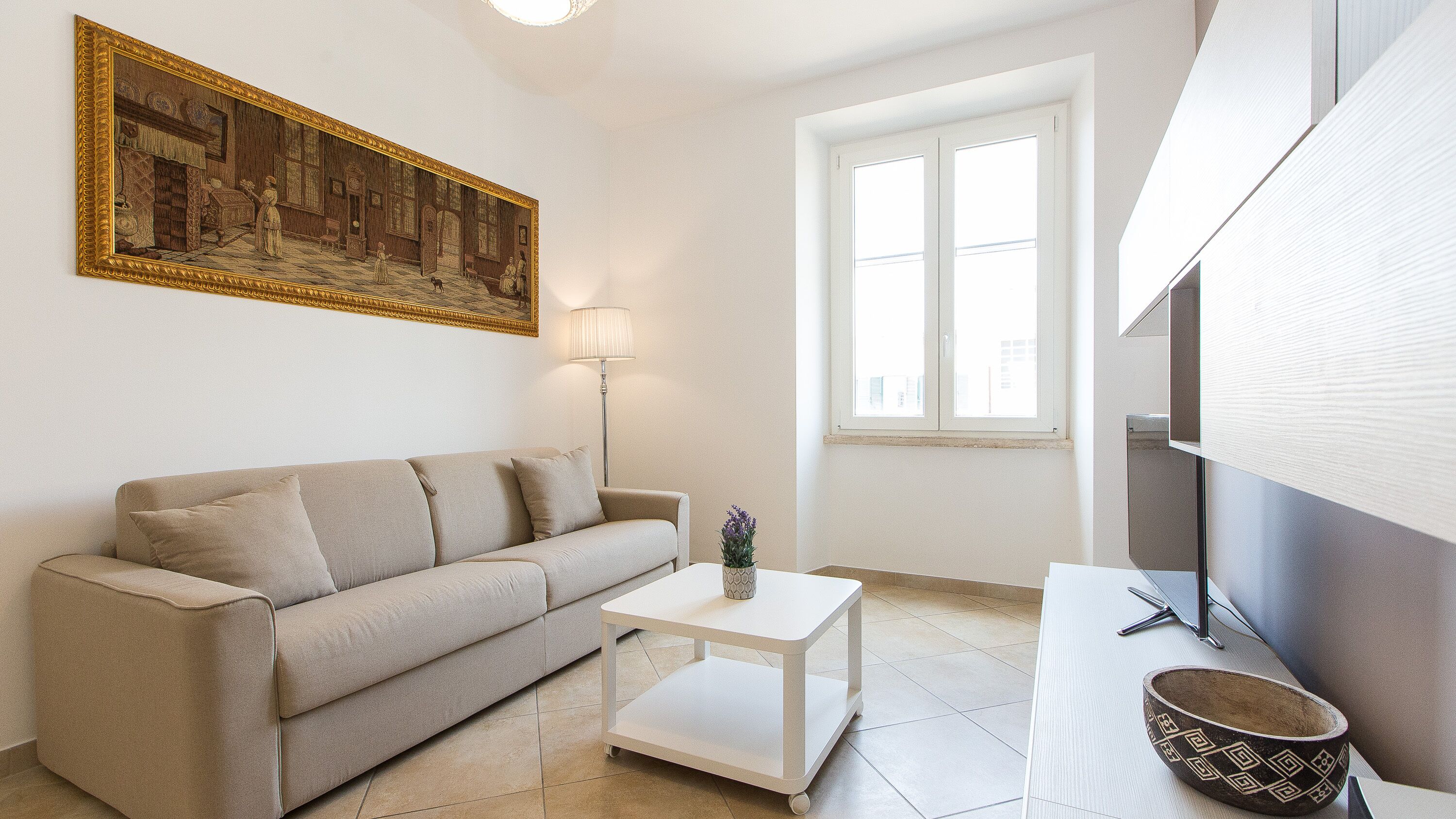 Een woonkamer met een bank, een salontafel, een lamp, een schilderij en een raam.