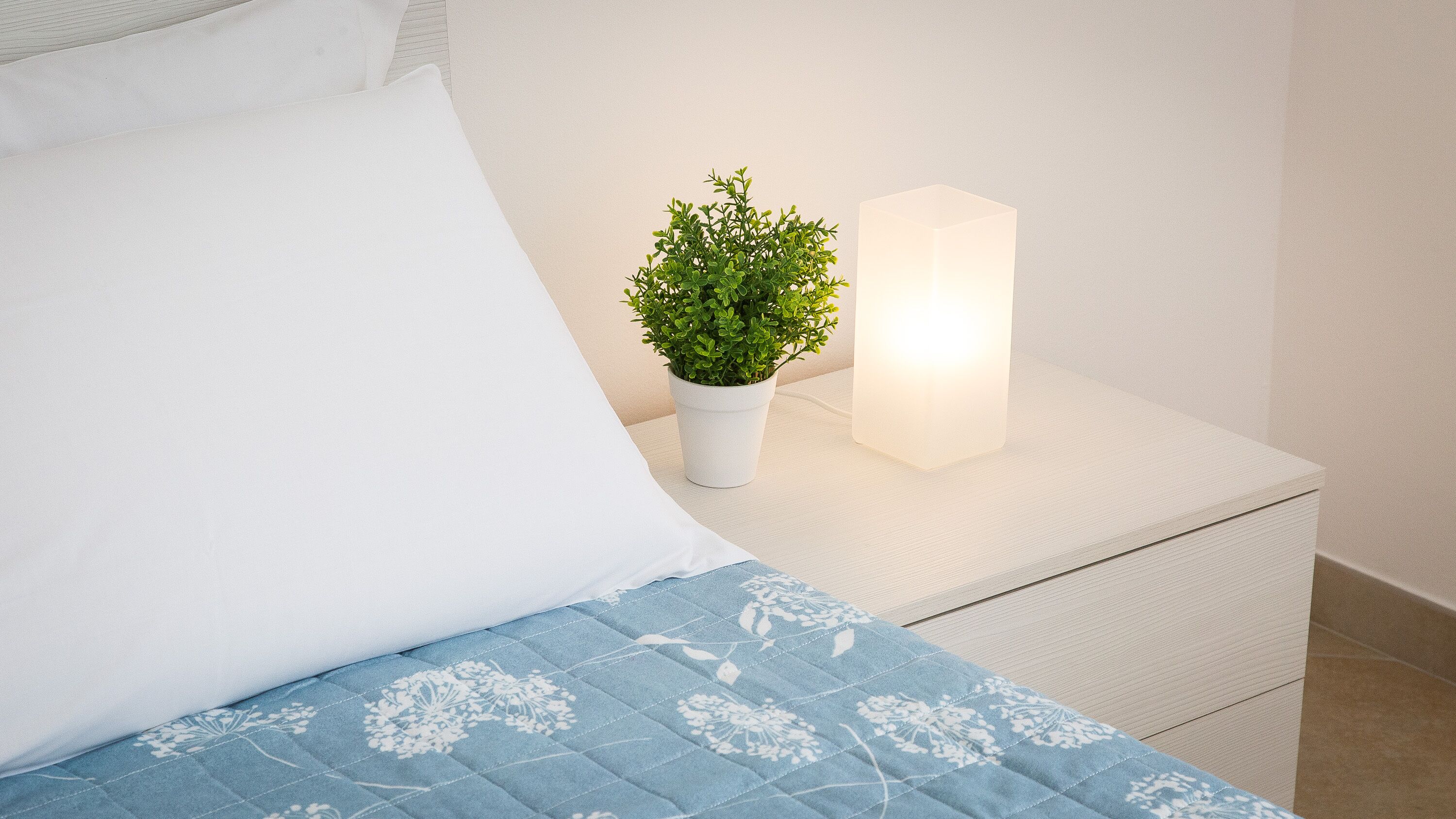 Een net opgemaakt bed met een blauwe bloemenbedekking, een wit kussen, een witte nachtkastje met een potplant en een brandende kaars.