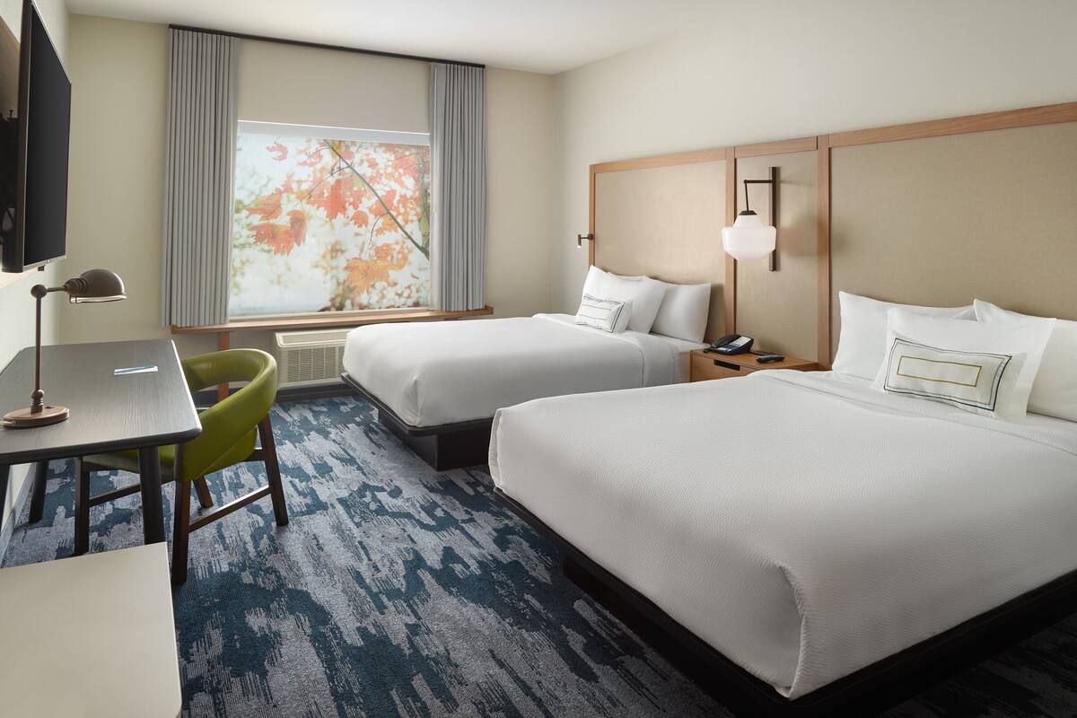 A hotel room with two beds, a desk, a chair, a TV, and a large window with curtains.