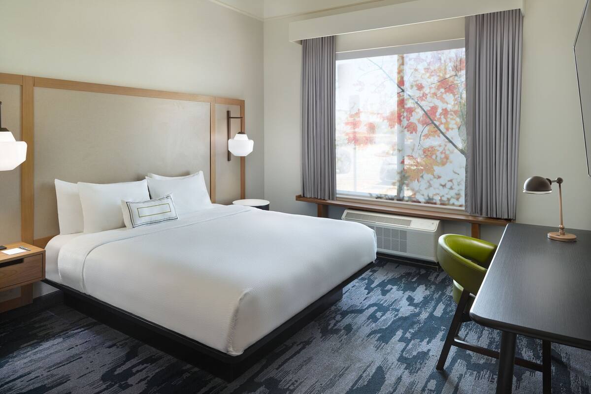 A hotel room with a large bed, a desk, a chair, a window with curtains, and a lamp.