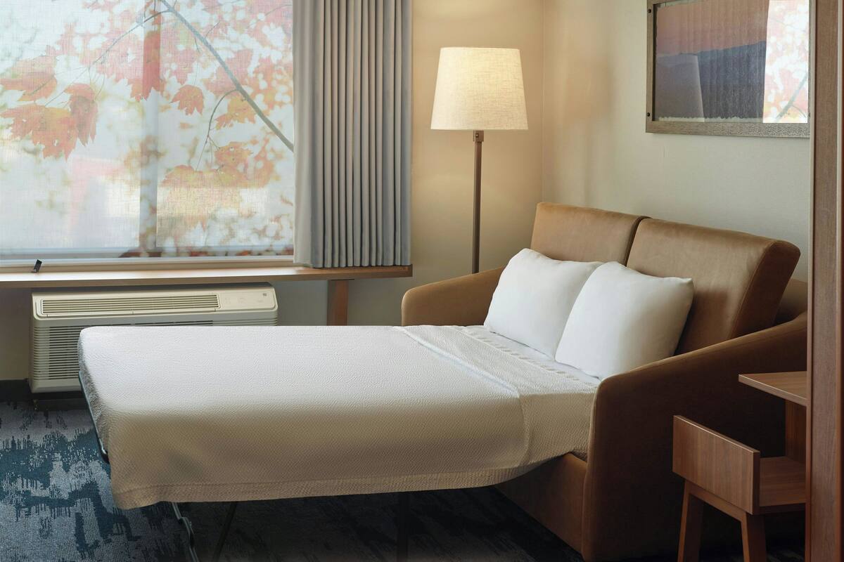 A hotel room with a bed, a bedside table, a lamp, and a window with a view of trees.