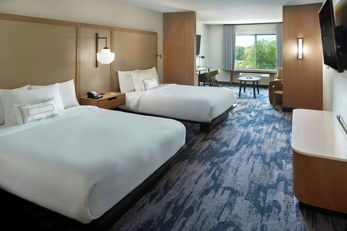 A hotel room with two beds, a desk, a chair, a TV, and a window with a view of trees.