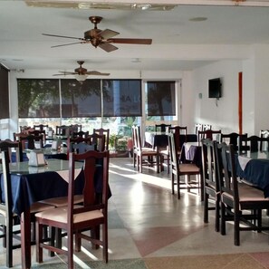 Un restaurant amb diverses taules i cadires, ventiladors de sostre i grans finestres.