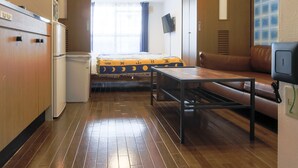 A compact room with a wooden floor, a bed with a yellow and blue bedspread, a brown leather sofa, a small table, and a kitchenette area. - Grand Court Namba 402 (Osaka)