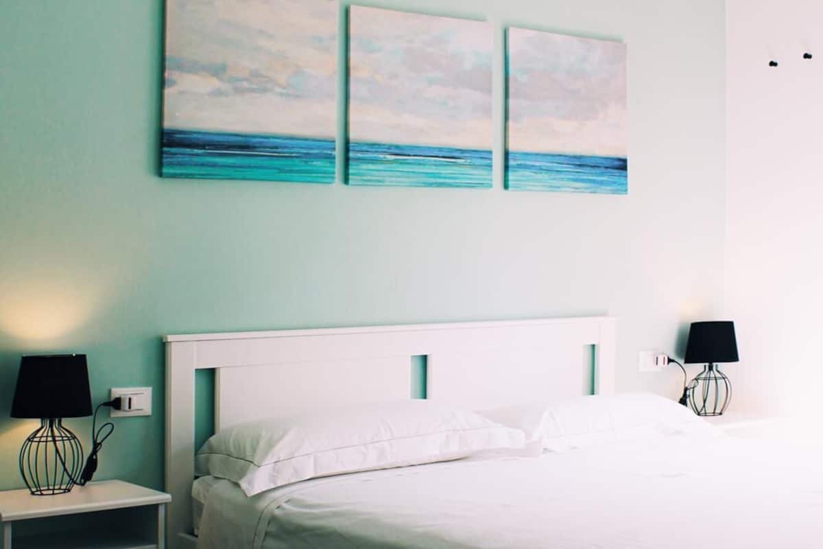 A neatly made bed with white linens, a white headboard, and a nightstand with a lamp. There are three wall-mounted paintings above the bed.