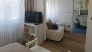 Una habitación pequeña y luminosa, con una cama, un televisor sobre un mueble bajo, un escritorio blanco y una silla. Se observa una pequeña cocina americana al fondo.