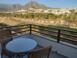 A balcony with wicker chairs and a glass table, overlooking a town with buildings and palm trees, and mountains in the background. - South Juan palms (Playa de la Américas)