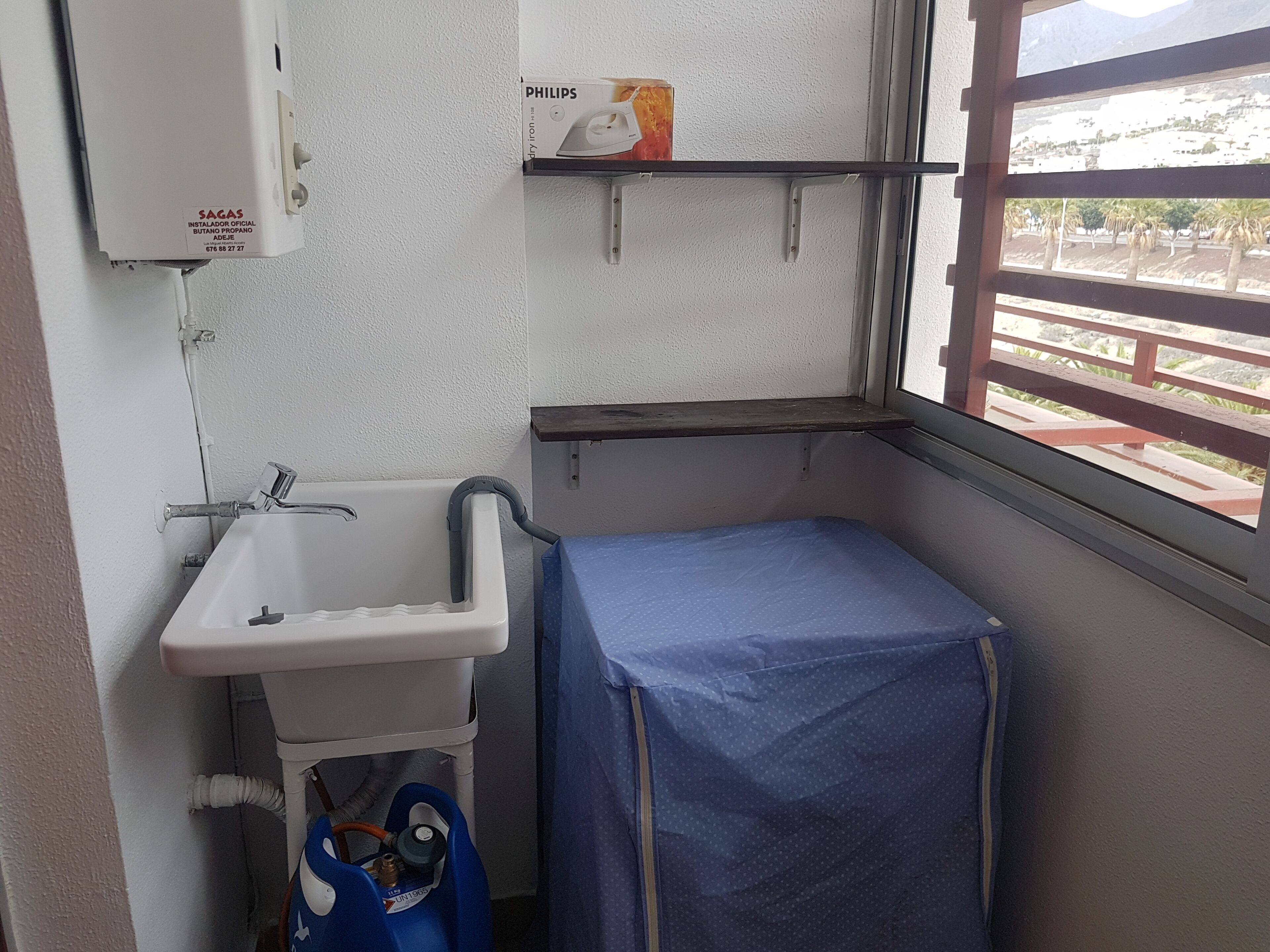 A small bathroom with a sink, a shelf with a Philips brand iron, and a window with a view of a building and palm trees.