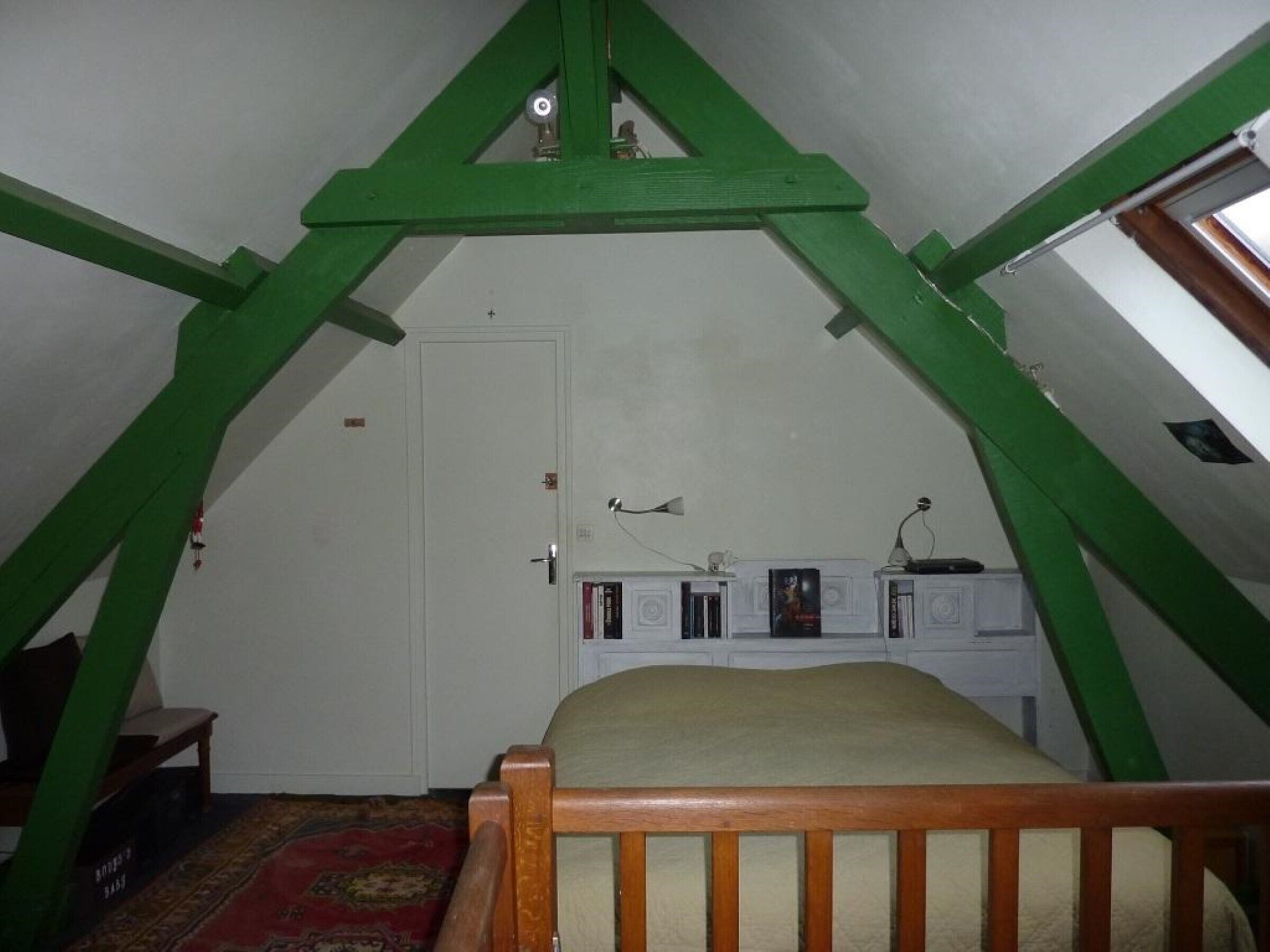 Une chambre avec un plafond en pente, un lit, un petit bureau avec une lampe et une porte.