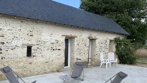 Een gebouw van steen met een pannendak, een kleine deur en een raam. Buiten staan vier lounge fauteuils en een tafel.