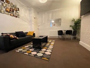 A modern living room with a black sofa, a coffee table, and a patterned rug. There is a dining area with a table and chairs.