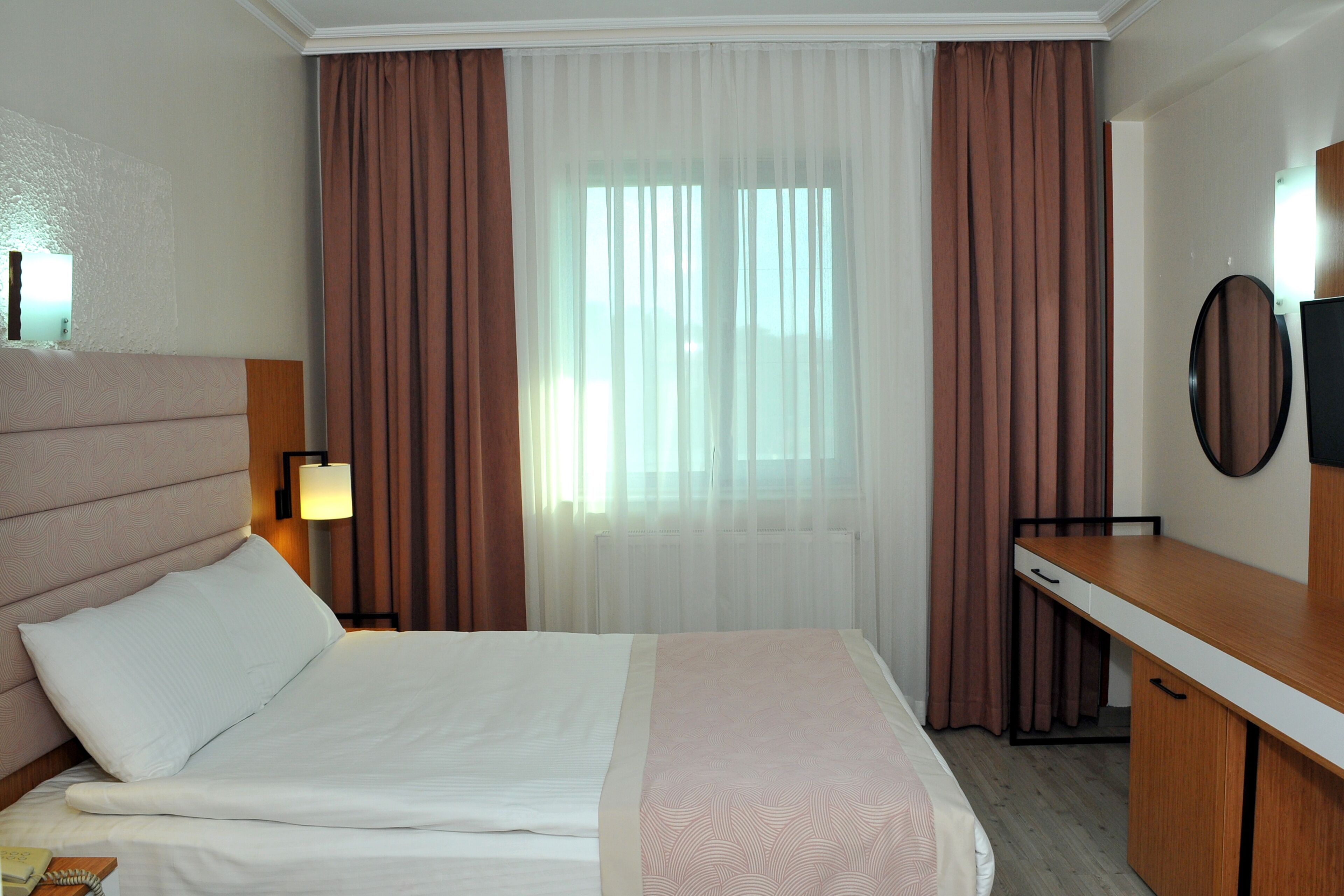 A hotel room with a bed, a desk, a chair, a lamp, and a mirror.