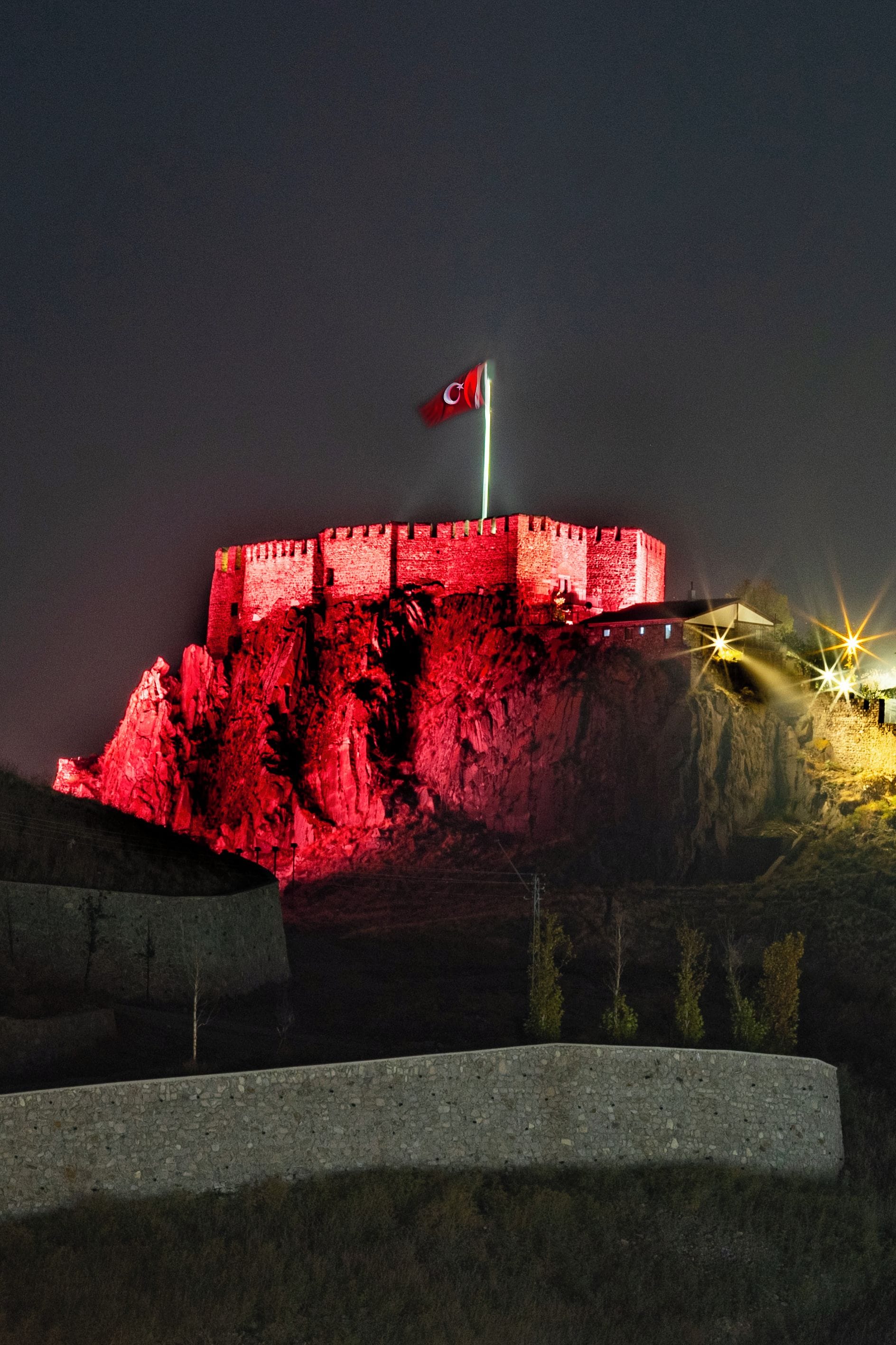 Une vaste structure imposante, semblable à une forteresse, illuminée et surmontée d’un drapeau, entourée d’un mur de pierre et d’une zone herbeuse.