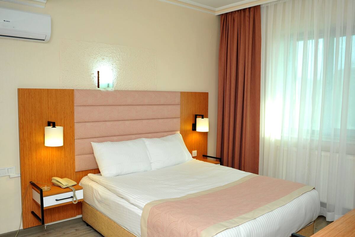 A hotel room with a large bed, a bedside table with a lamp, a telephone, and a wall-mounted air conditioner.