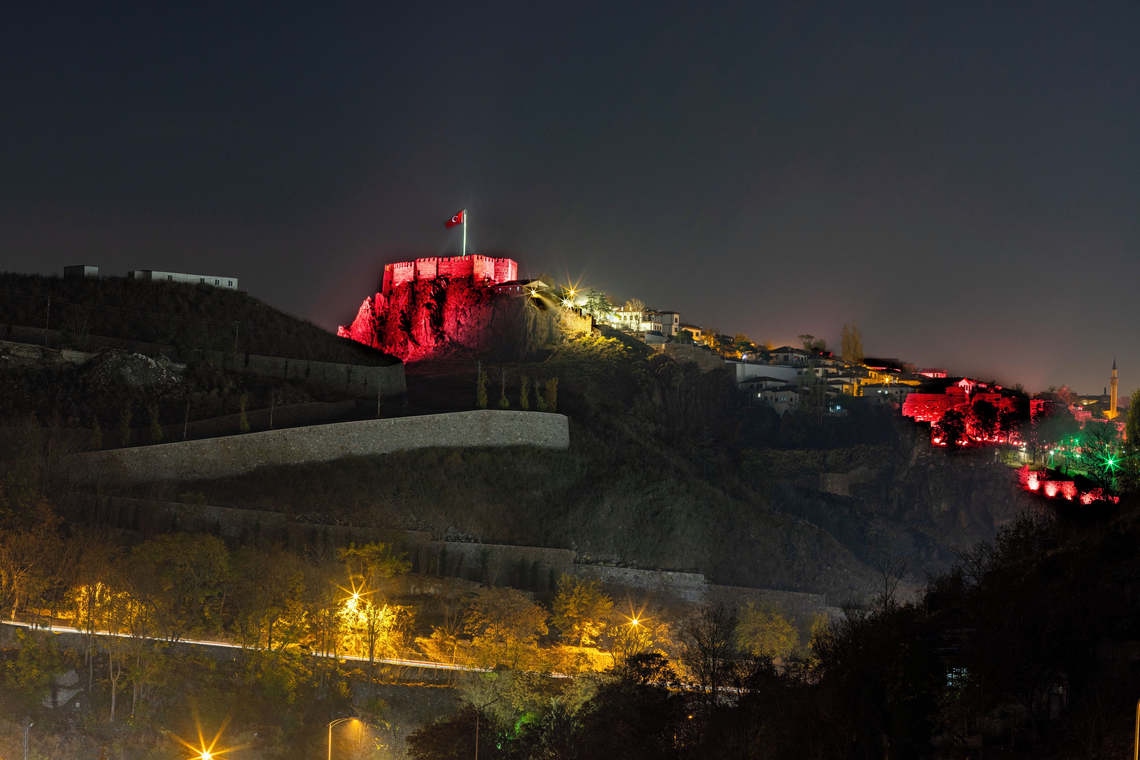 Una estructura fortificada con una bandera en la parte superior, rodeada por un paisaje montañoso con senderos iluminados y edificios.