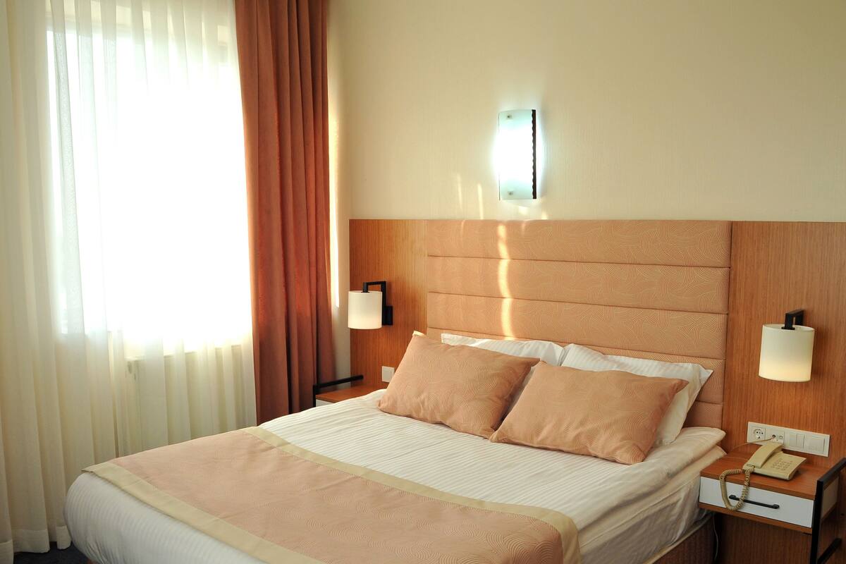 A hotel room with a bed, two bedside lamps, a nightstand with a telephone, and a window with curtains.