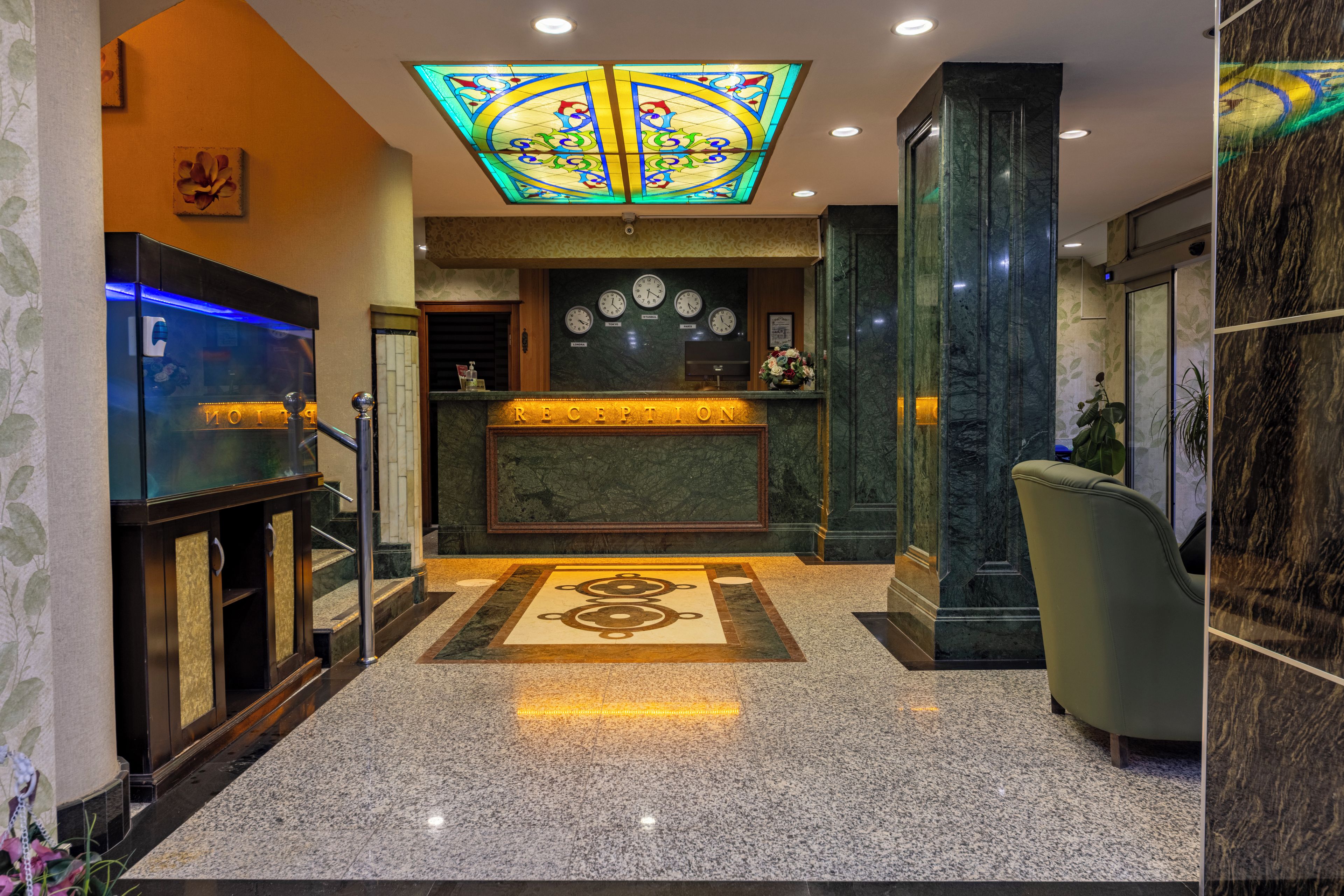 A hotel lobby with a reception desk, a large stained glass window, and a clock displaying multiple time zones.