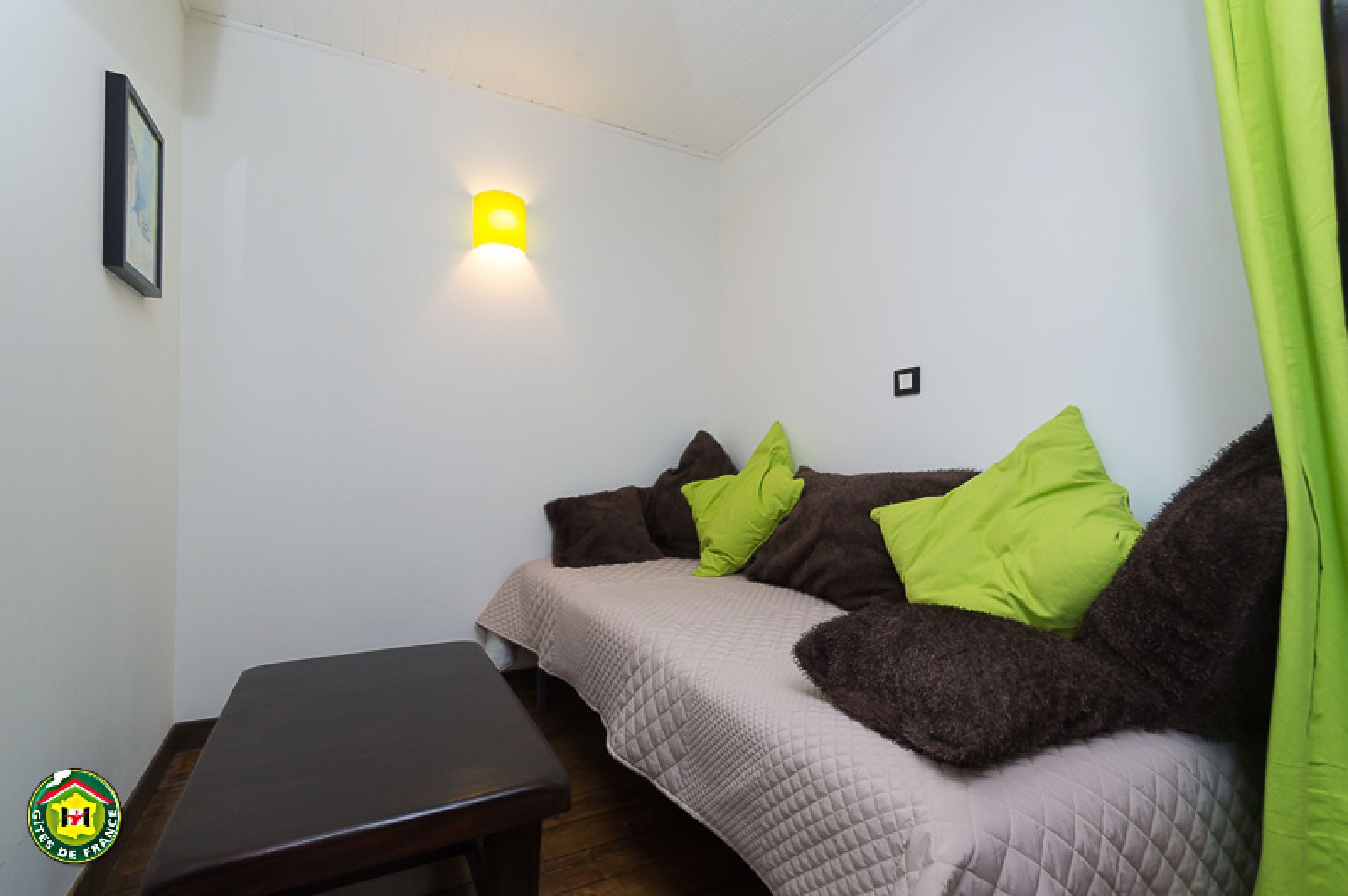 A small, cozy room with a bed, a bench, and a wall-mounted light.