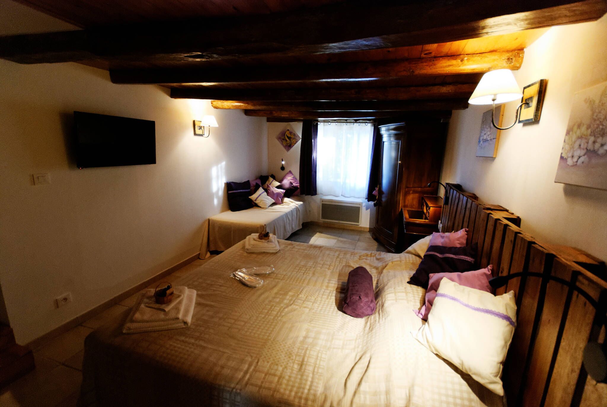 Une chambre à coucher avec un grand lit, un lit plus petit, une tête de lit en bois, un téléviseur à écran plat et une fenêtre avec des rideaux.
