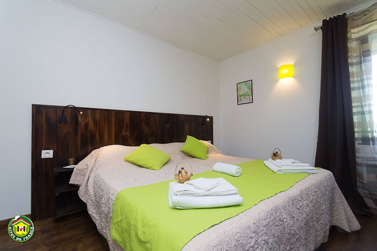 Uma cama bem arrumada com almofadas verdes, uma mesinha de cabeceira, uma luminária fixada na parede e um quadro emoldurado.