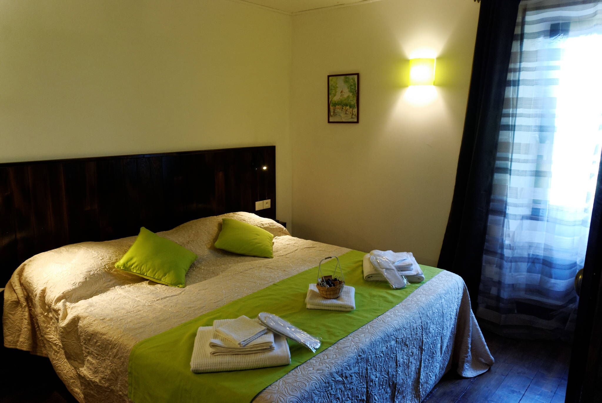 A hotel room with a bed, green pillows, a green bedspread, and a bedside table with towels and a basket.