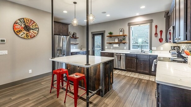 A modern kitchen with a central island, red bar stools, pendant lighting, and a large clock on the wall.