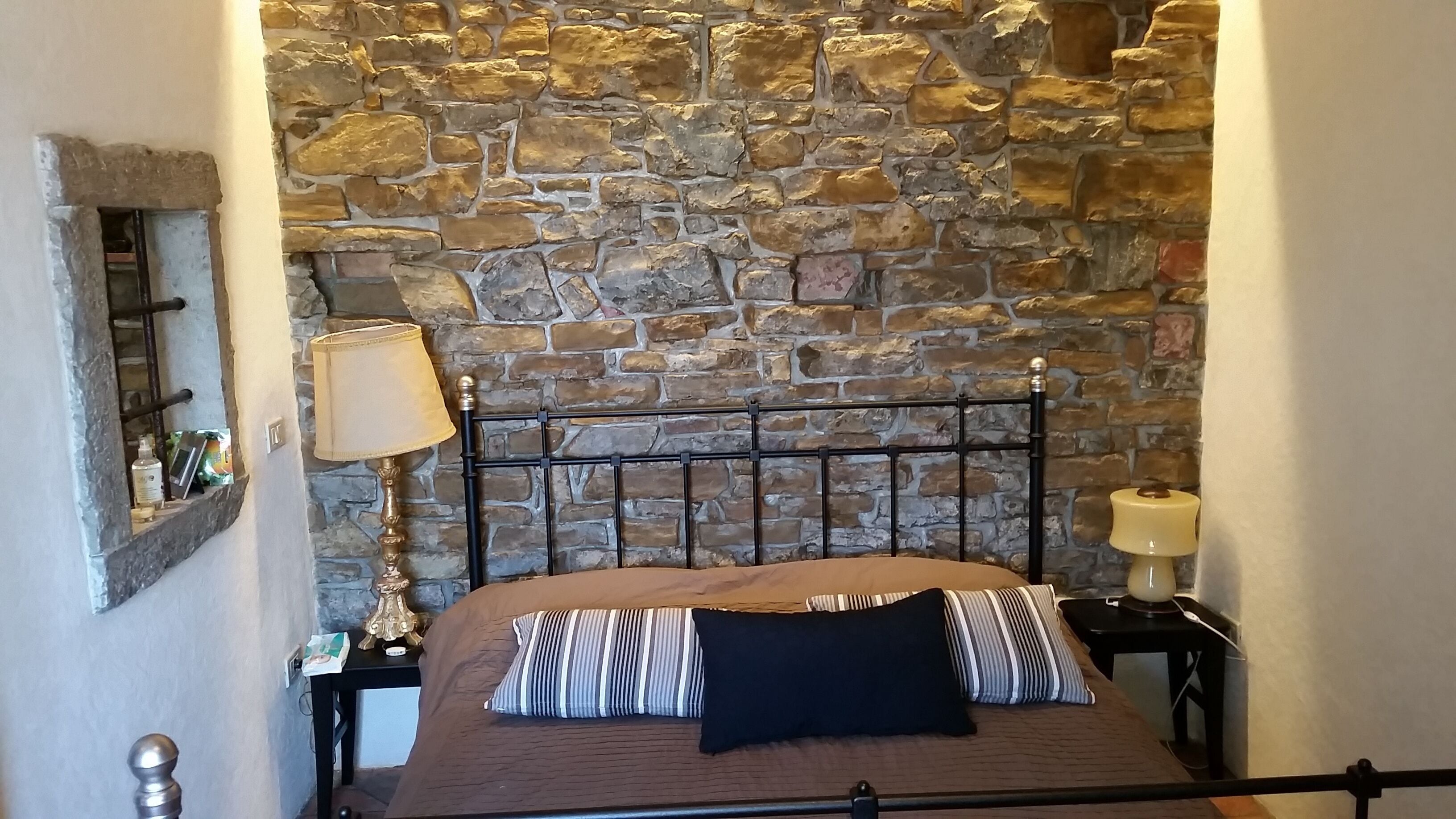 Un dormitorio con pared de piedra, estructura de cama de metal, mesita de noche con lámpara y otro mueble auxiliar con lámpara.