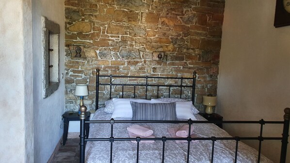 A bedroom with a stone wall, a metal bed frame, a nightstand with a lamp, and a bedside table with a lamp. - Monfort Apartments (Piran)