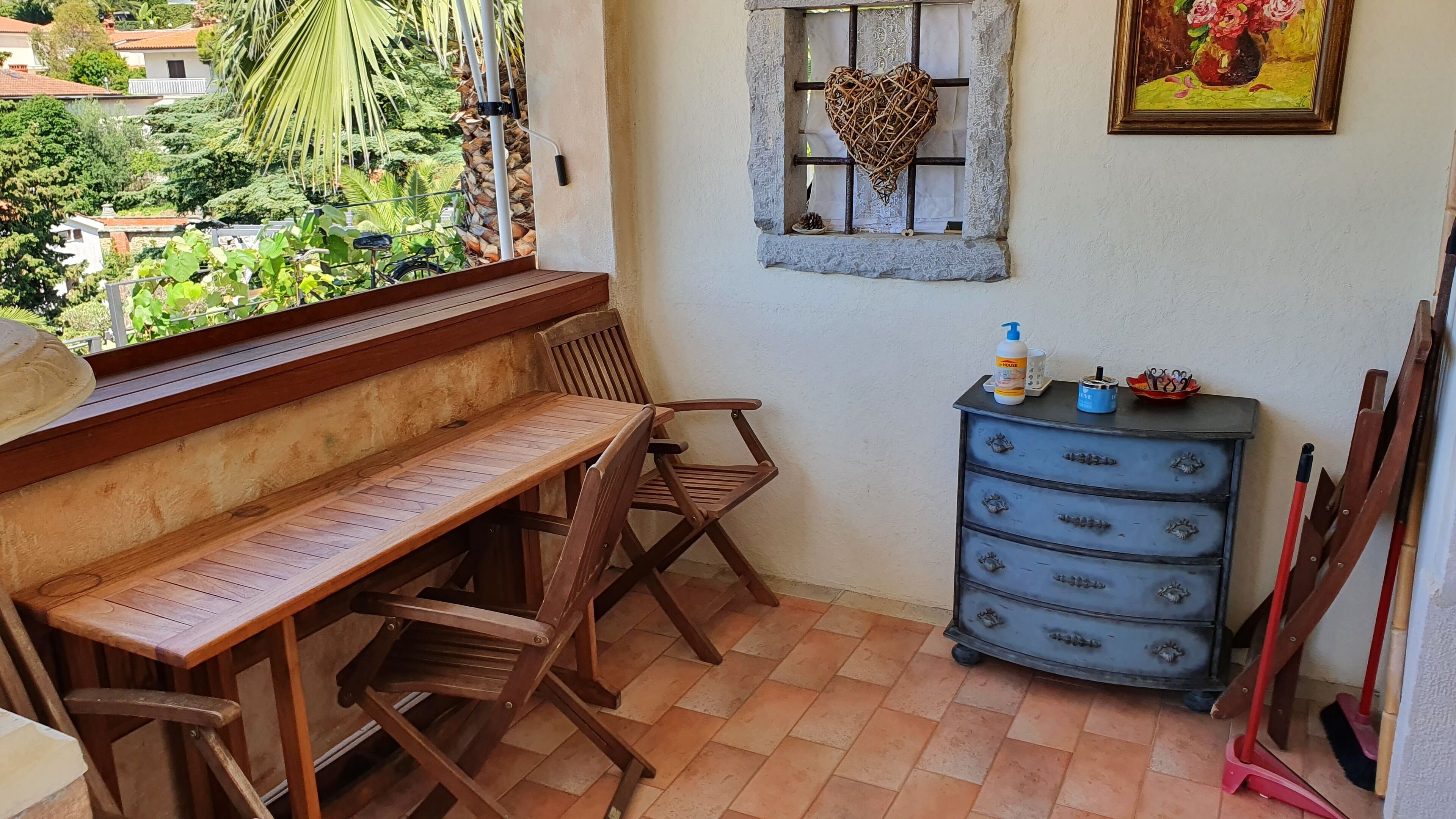 Un balcón con una mesa y sillas de madera, un pequeño aparador con cajones, una ventana decorada con un corazón y un cuadro en la pared.