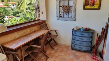 Un balcón con una mesa y sillas de madera, un pequeño aparador con cajones, una ventana decorada con un corazón y un cuadro en la pared.