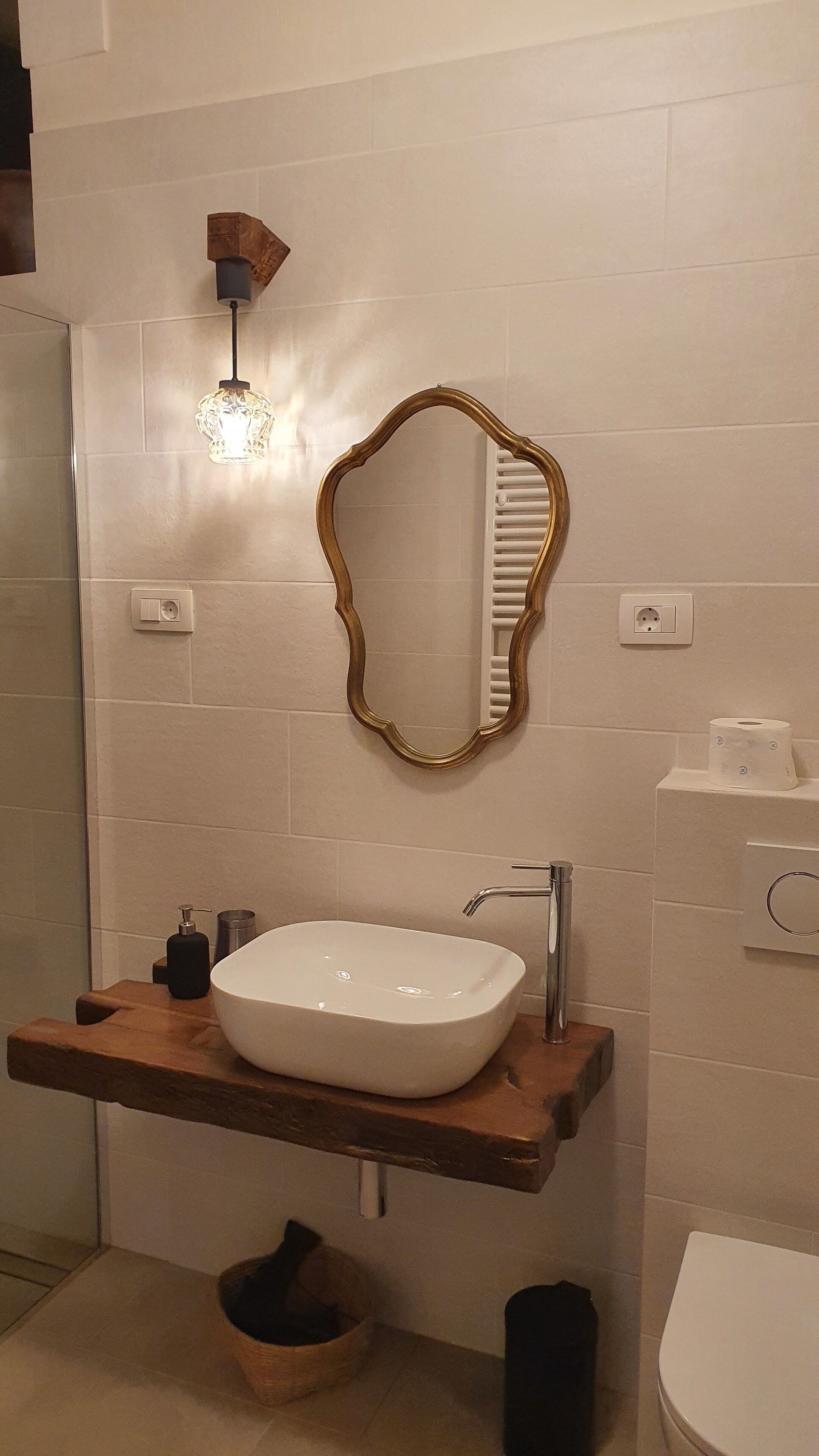A bathroom with a white sink, a wooden vanity, a gold-framed mirror, a wall-mounted light, and a toilet.