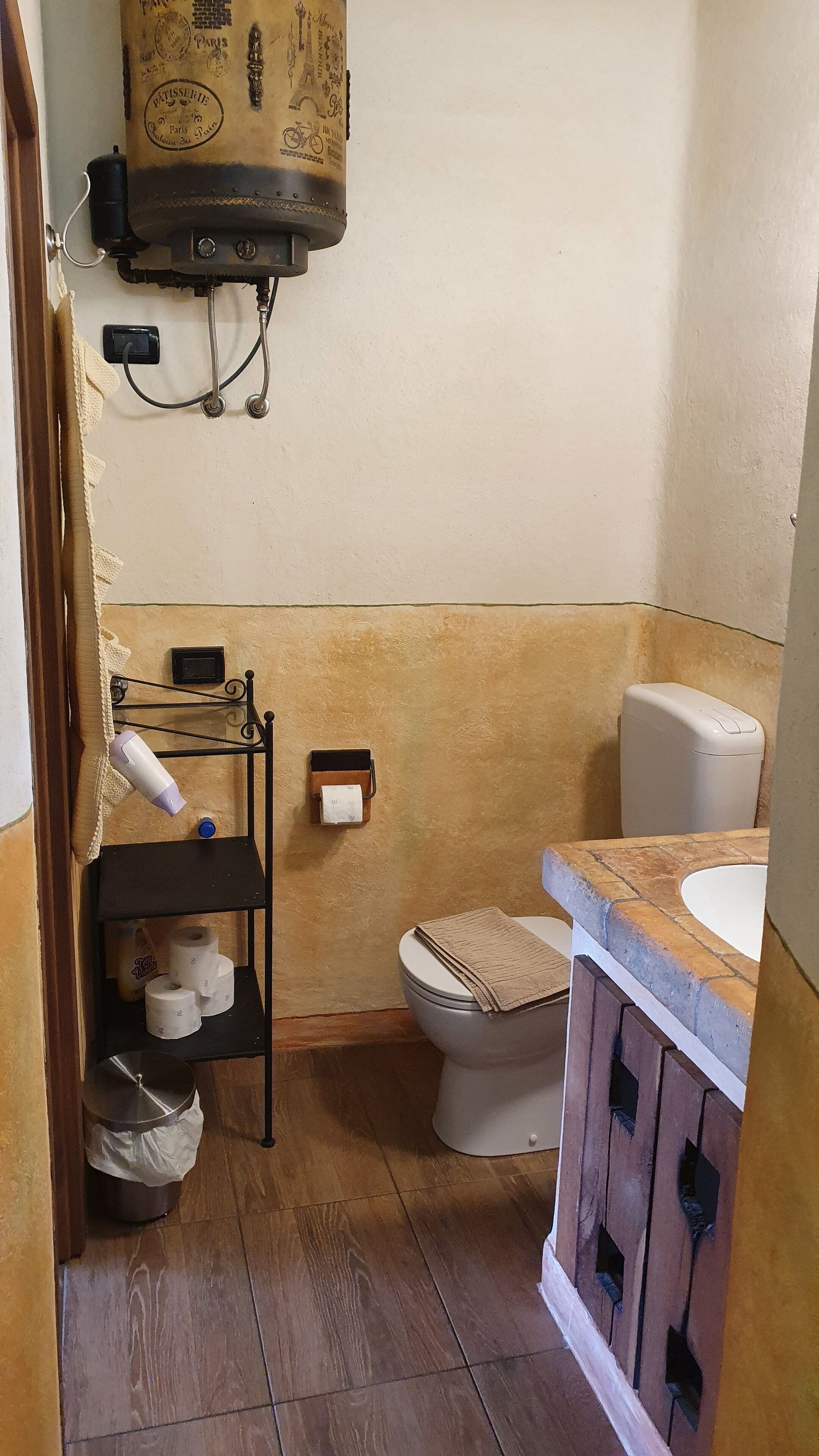A compact bathroom with a toilet, a sink, a towel rack, and a shelf with toiletries.