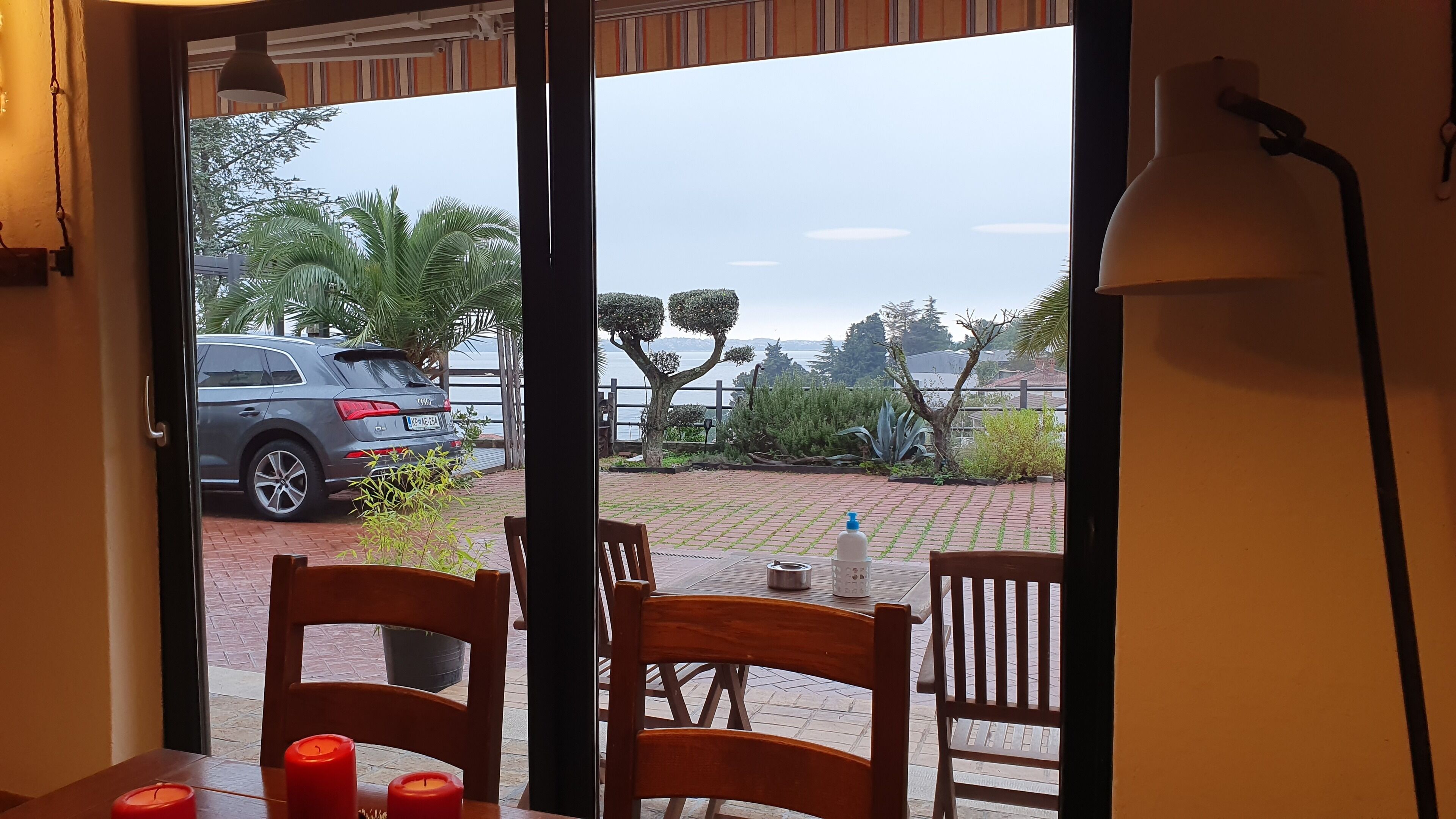 Blick von einem Zimmer mit Tisch und Stühlen, ein Fenster mit Blick auf ein parkendes Auto, eine Flasche und eine Kerze.