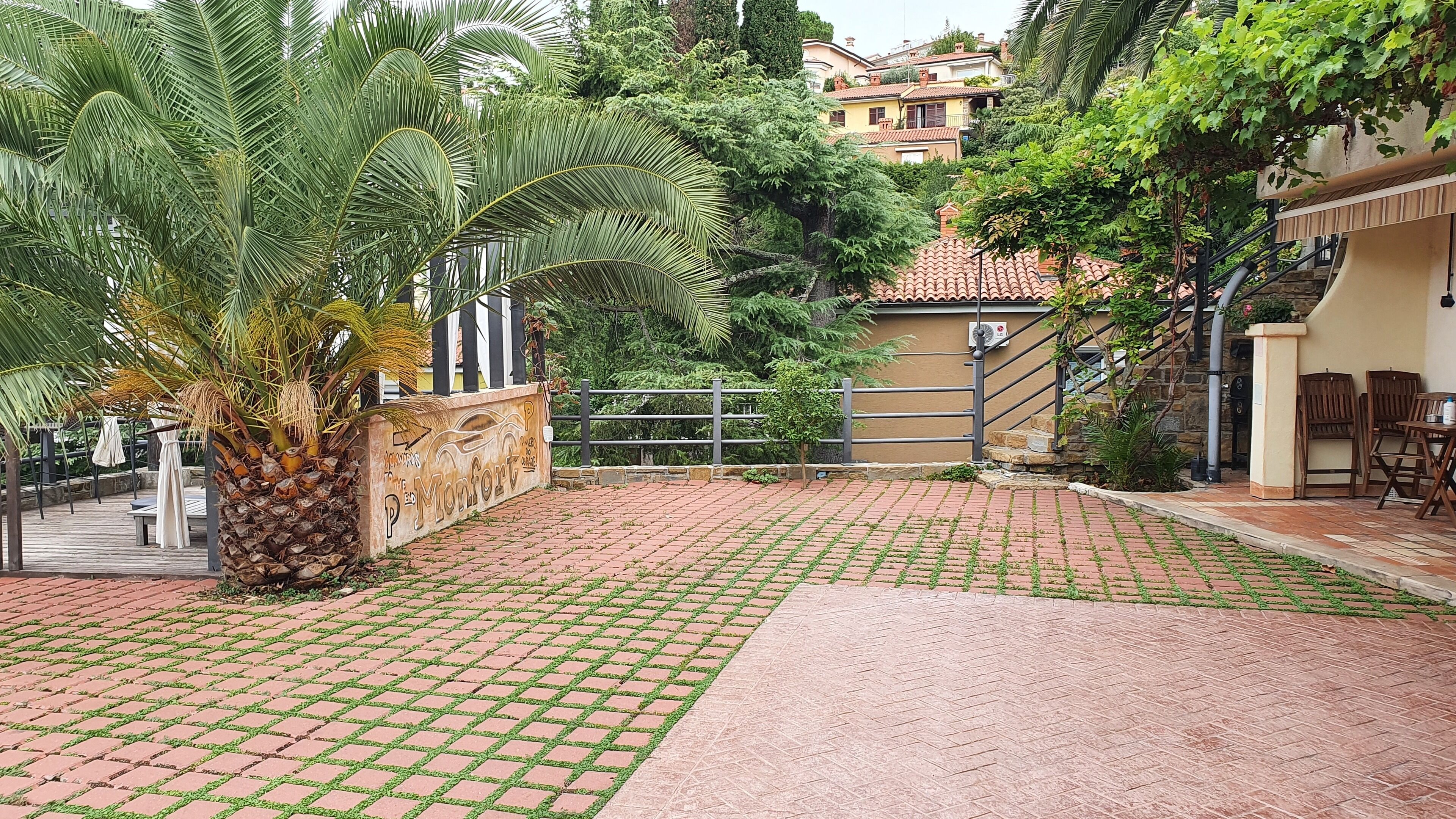 Pátio pavimentado com palmeiras, muro de pedra com grafite e escadaria que leva a um edifício com telhado em azulejos.