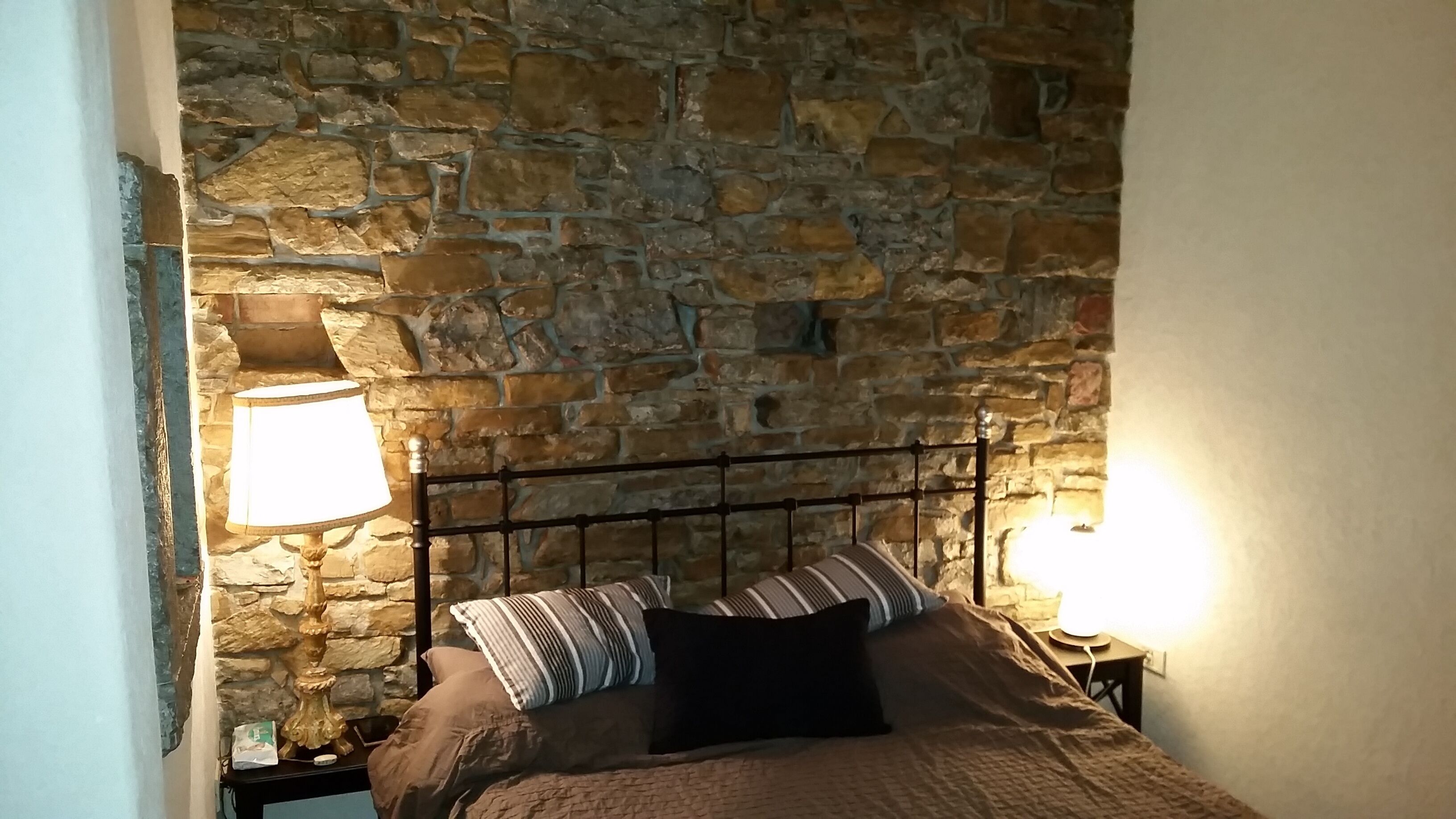 Cama com parede de pedra ao fundo, duas luminárias de cabeceira e um travesseiro listrado.