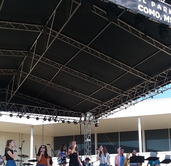Musicians perform on a stage with instruments and microphones under a canopy.