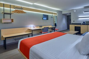 Una habitación de hotel moderna con una cama grande, un escritorio con silla, una kitchenette pequeña y un televisor montado en la pared.