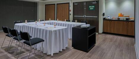A conference room with a long table, chairs, a projector, and a kitchenette area with a sign for the exit.
