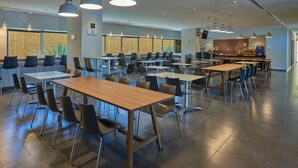 Une cafétéria spacieuse, dotée de nombreuses tables et chaises, de grandes fenêtres ornées de plantes et d’un espace cuisine moderne.