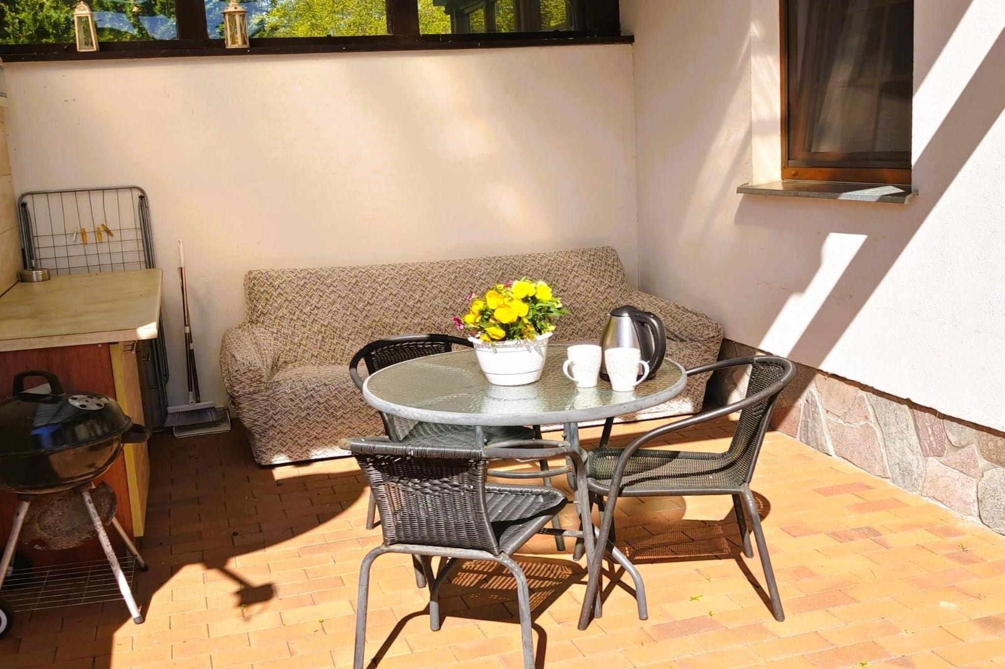 Un espace patio comprenant une table ronde, deux chaises, un canapé, un barbecue et une fenêtre donnant sur un paysage verdoyant.