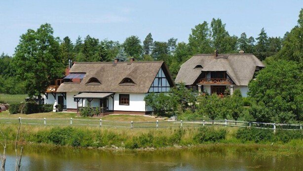 A traditional thatched-roof house with a white exterior, situated by a body of water, surrounded by greenery. - Apartment only 20 m from  Lake in Rekowo (Rekowo)