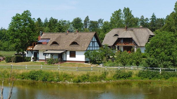 A traditional thatched-roof house with a white exterior, situated by a body of water, surrounded by lush greenery.