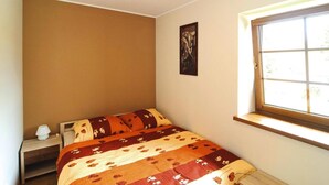 A single bed with a floral-patterned bedspread, a bedside table with a lamp, and a window with a view of greenery.