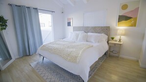 Un dormitorio con cama, mesita de noche, lámpara, ventana con persianas y un cuadro en la pared.