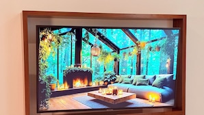 Een lijst met een televisie waarop een gezellige kamer te zien is met een open haard, bank en potplanten.